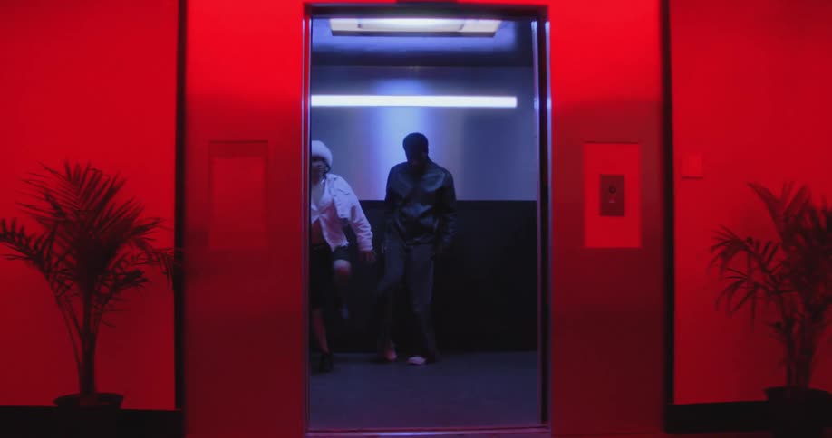 An elevator door opens in a surreal, red-lit hallway, revealing a stylish couple performing an intense and energetic dance routine inside