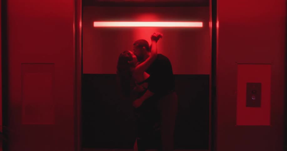 A couple passionately kisses inside an elevator bathed in sensual red light as the camera pulls back and the doors close