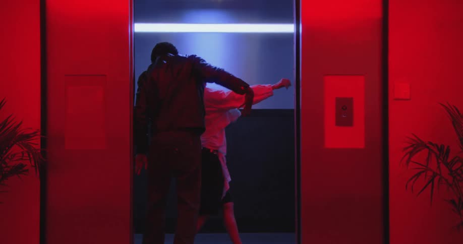 A stylish man and woman perform an energetic hip-hop dance duet inside an elevator illuminated by vibrant red and blue neon lights