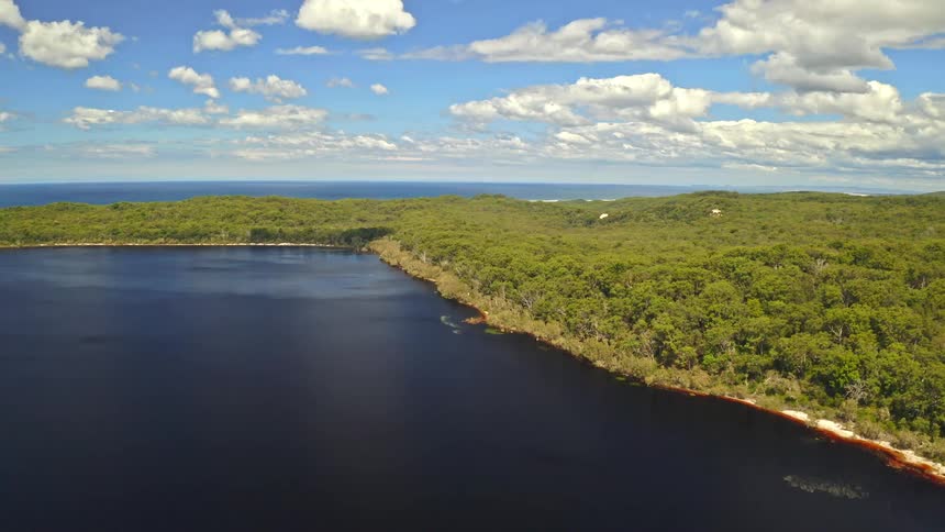 A beautiful aerial shot captures the stunning landscape where a dark lake meets a dense green forest, with the vast ocean in the background