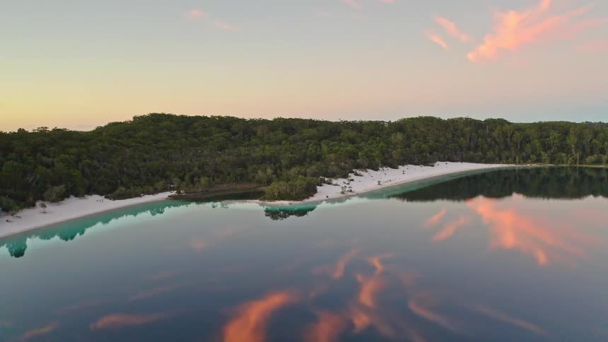 A serene aerial view descends over a pristine lake with a white sand shoreline, reflecting the beautiful colors of the sunset clouds on its mirror-like surface