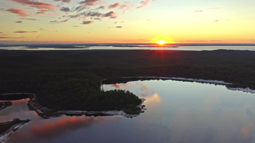 Stunning aerial footage captures the sun setting over a vast forest and a tranquil lake reflecting the golden sky