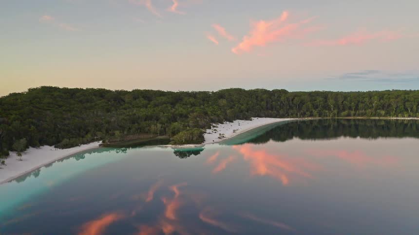 Stunning aerial footage captures the serene beauty of a freshwater lake with a white sand beach at sunset, with colorful clouds reflected in the still water