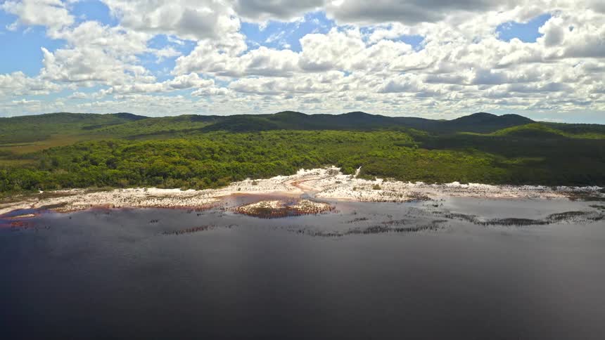 A wide aerial shot captures the vast landscape of a dark water lake meeting a sandy shoreline backed by a dense green forest and rolling hills under a cloudy sky