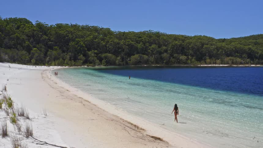 A woman in a bikini walks along the pristine white sand shore of a beautiful blue lake on a sunny day