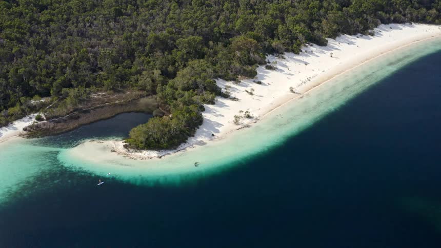 An incredible aerial view tilts down over a breathtaking lake, revealing its white sand beach and crystal-clear turquoise waters bordered by a lush forest