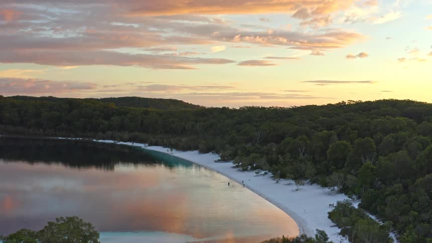 A breathtaking aerial view captures a pristine white sand beach and tranquil lake surrounded by a lush forest during a vibrant sunset