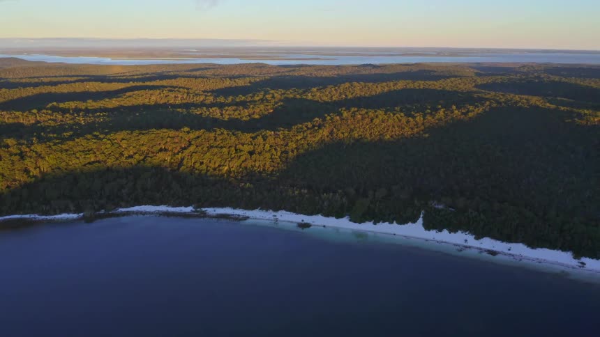 A stunning aerial view pulls back from a white sand beach and deep blue lake to reveal a vast, dense forest glowing in the golden light of sunrise