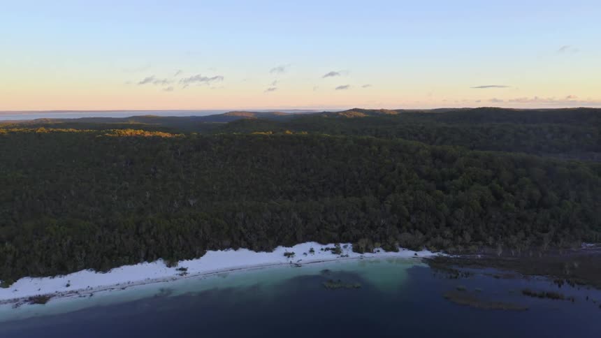 Stunning aerial pullback reveals a white sand beach and tranquil lake surrounded by a dense forest at sunrise