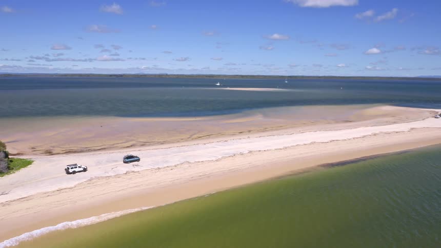 A stunning aerial view pans across a long sand spit where off-road vehicles are parked on the beach next to the vast ocean
