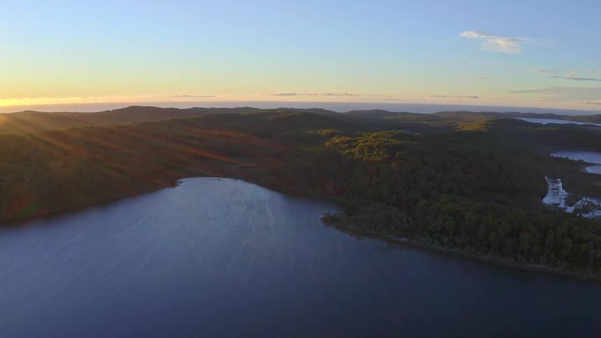 A majestic aerial view of a serene lake and rolling, forested hills illuminated by the warm, golden glow of a setting sun