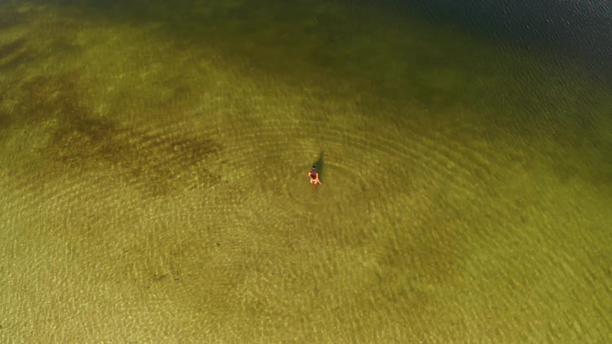 An aerial view slowly tilts down to a top-down shot of a solitary woman standing in the clear, rippling waters of a beautiful lake