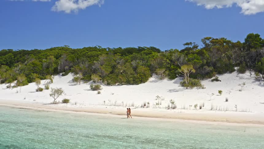 An aerial view follows a young couple walking along the pristine white sand shore of a beautiful freshwater lake on a sunny island vacation