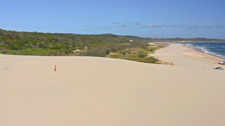 A woman in a bikini walks across a vast sand dune, revealing a beautiful sunny beach and coastline