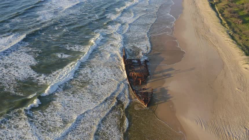 A stunning top-down aerial shot of a rusty shipwreck on a sandy beach pulls back to reveal the vast ocean and endless coastline at sunset