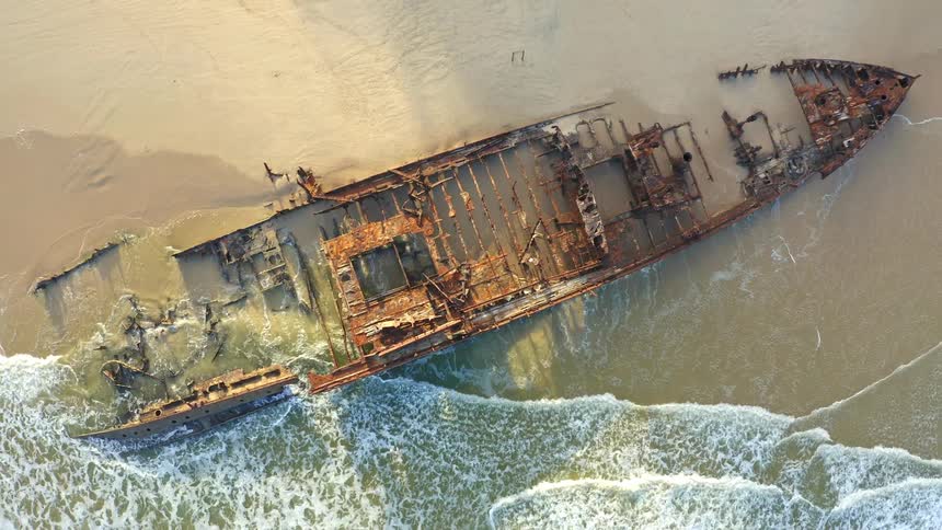 A dramatic top-down aerial view zooms out from a large, rusty shipwreck being reclaimed by the ocean on a sandy beach