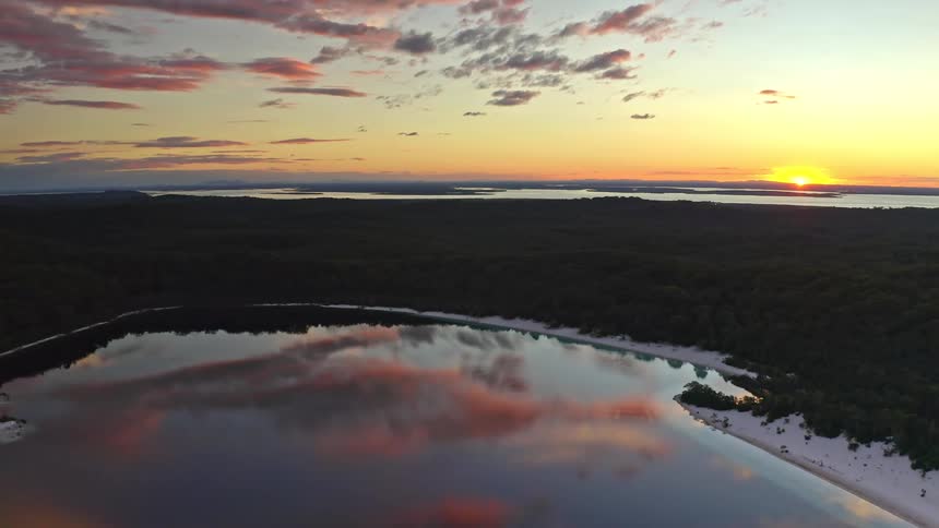 A cinemagraph-style aerial view shows a tranquil lake reflecting the beautiful sunset clouds over a vast forest landscape