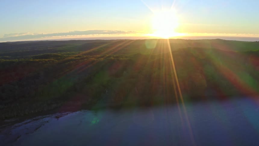 Slowly pulling back aerial shot reveals a stunning sunrise casting brilliant golden rays over a vast forested landscape and a calm lake