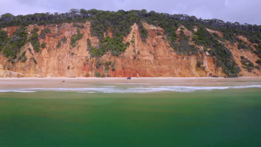 Drone pulls back from an off-road vehicle parked on a remote beach to reveal stunning red cliffs and an expansive green ocean