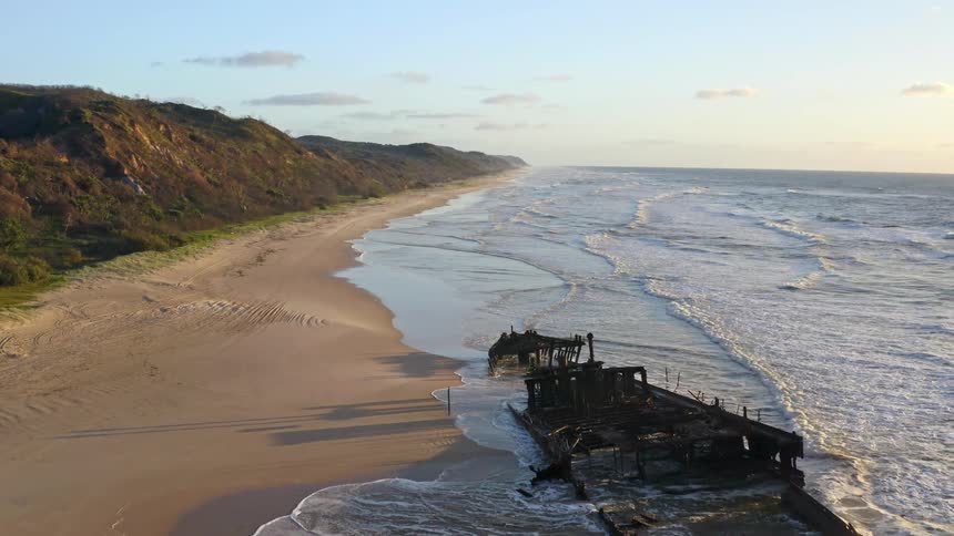 Aerial reveal of a rusted shipwreck beached on a long sandy coastline as gentle waves roll in