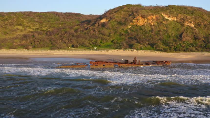 Aerial view slowly pans around a large, rusty shipwreck beached on a sandy shore with rolling waves and a green coastline in the background
