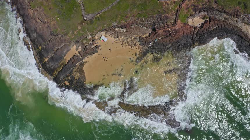 A static top-down aerial view shows powerful ocean waves crashing into a small, secluded sandy cove surrounded by rugged, rocky cliffs