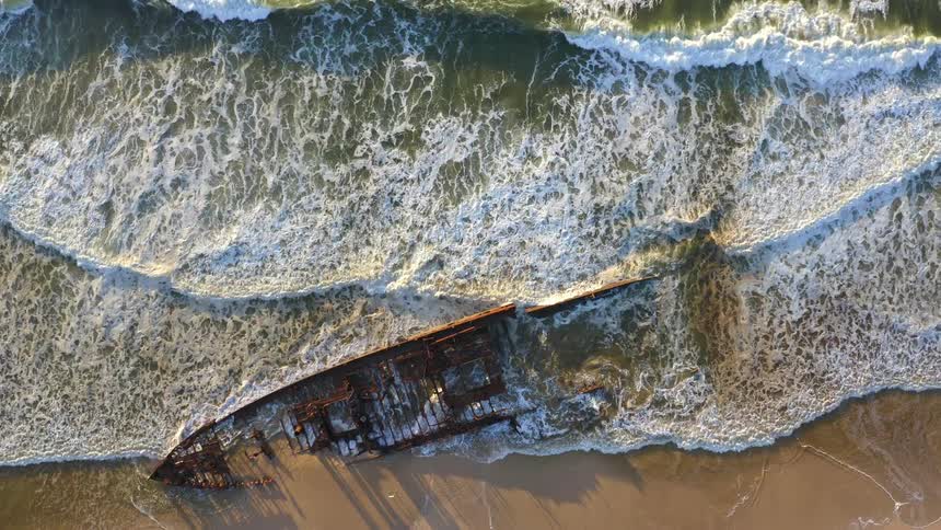 Static aerial top-down shot of a decaying shipwreck on a golden sand beach as foamy ocean waves crash ashore