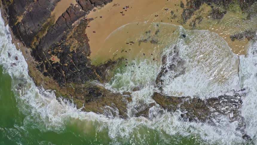 A stunning aerial ascent reveals people enjoying a secluded, rocky tide pool on a beautiful beach
