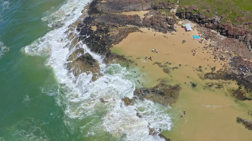 An aerial top-down view shows people enjoying a sunny day, swimming and relaxing in a picturesque rocky cove as ocean waves crash ashore