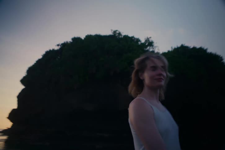 A free-spirited woman with blonde hair dances and spins in slow motion on a secluded beach at twilight