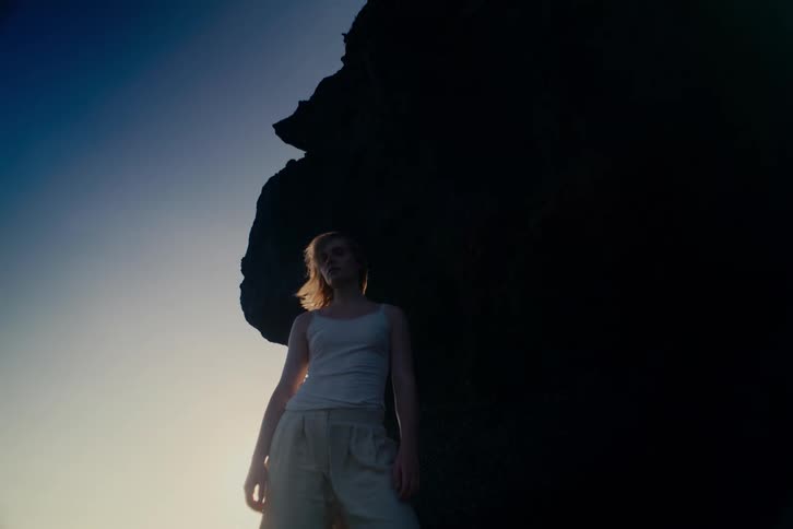 A thoughtful young person stands against a shadowy cliff, backlit by the soft glow of the sun