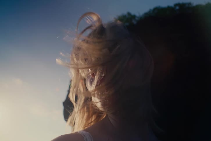 A beautiful blonde woman stands against the setting sun as the wind blows through her hair in slow motion