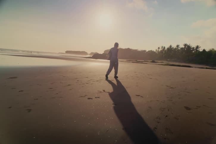 A surfer with a surfboard casts a long shadow while walking on a wide, reflective beach at sunset