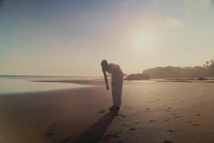 A woman in slow motion touches the sand and then begins a contemplative walk along a vast beach, backlit by the hazy setting sun