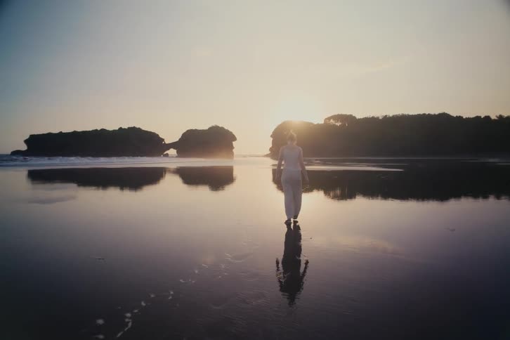 A lone woman dressed in white walks in slow motion across a vast, reflective shoreline during a golden sunset