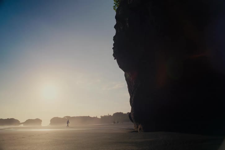 A lone silhouette walks along a vast, sandy beach next to a large rock formation as the bright sun hangs low in the sky