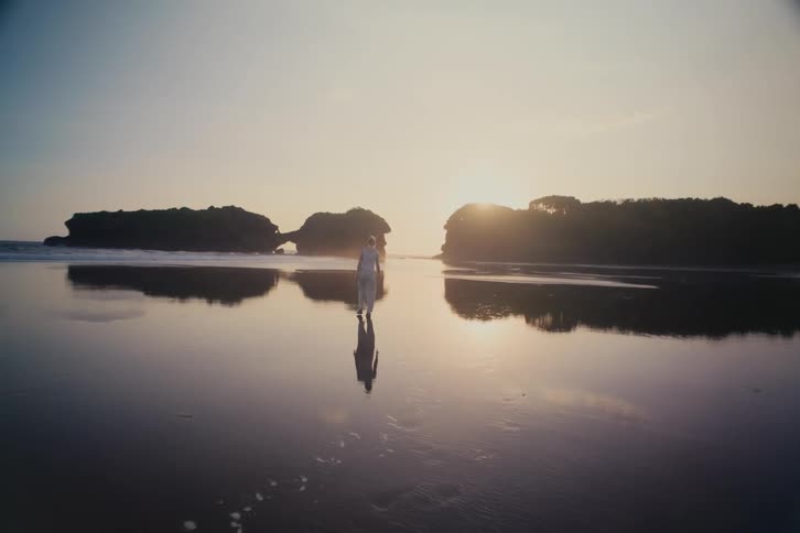 A woman in white takes a serene walk on a reflective beach during a golden sunset