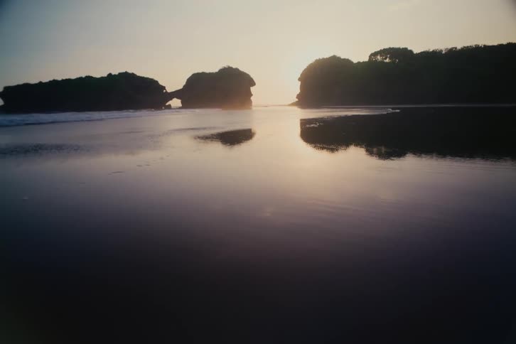 A serene sunset casts a warm glow over a calm beach, reflecting silhouetted rock formations in the gentle tide