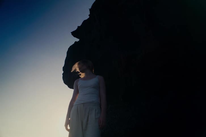 Low angle shot of a thoughtful woman with windblown hair standing by a massive rock as the sun flares behind her at golden hour