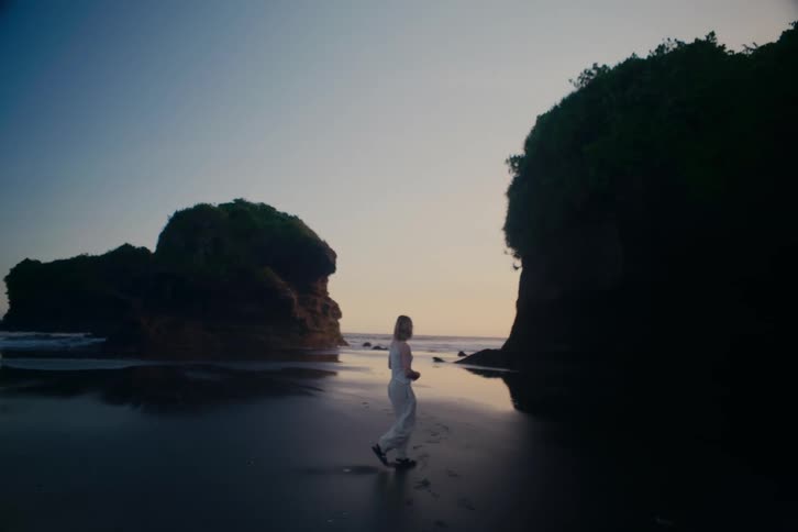 A woman in white walks on a serene black sand beach with dramatic rock formations during a beautiful sunset