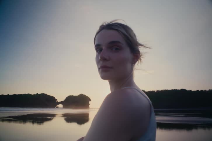 A serene young woman looks directly into the camera on a tranquil beach at sunrise, with golden light reflecting on the water