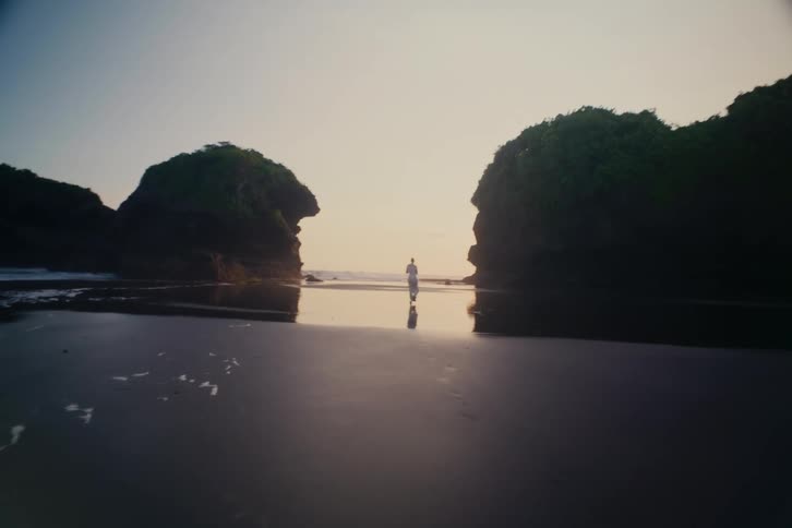 A person in white walks across a reflective wet sand beach towards the ocean between two large rock formations at sunset