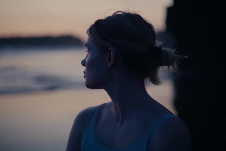 A thoughtful woman with her hair tied back gazes out at the tranquil water during a beautiful twilight glow