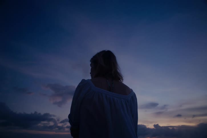 A still shot captures a woman's silhouette from behind as she contemplates the deep blue twilight sky after sunset