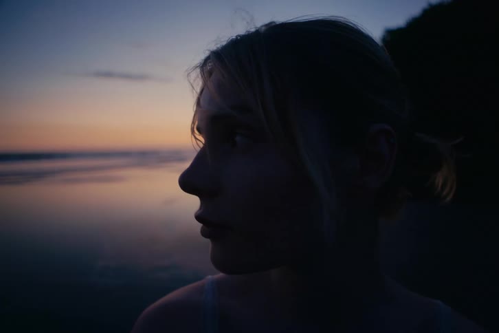 A close up shot of a pensive young woman's face in silhouette as she watches the sunset over the ocean