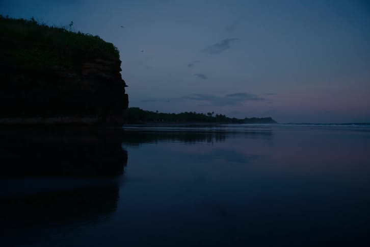 A still and reflective ocean mirrors a silhouetted cliff during the quiet moments of twilight