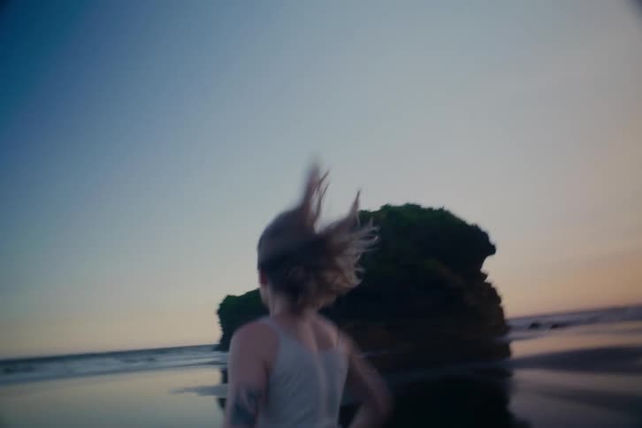 A young woman with blonde hair runs in slow motion along a secluded, wet sand beach between towering rock formations at dusk