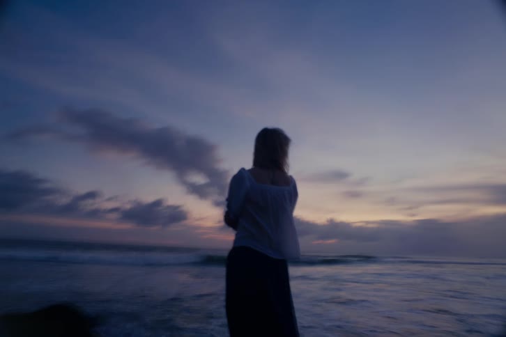 A dreamy shot comes into focus on a woman standing alone on the beach at twilight, watching the ocean waves
