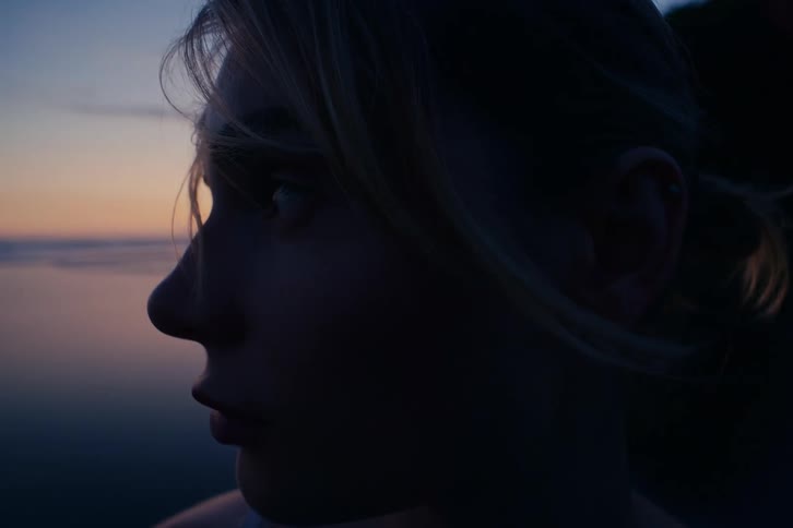 A thoughtful young woman's face is silhouetted in profile as she watches the sunset over the water