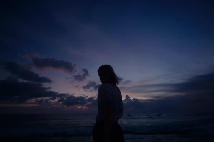 A woman stands silhouetted against a beautiful purple and blue twilight sky while looking out at the ocean